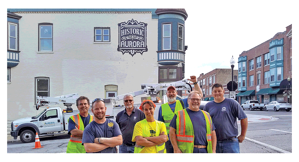 The new mural on Benton at LaSalle in downtown Aurora is a team collaboration. Team members stand in front of the new sign on the side of the former Pierpont Grocery. Look for more public art coming soon to walls and utility boxes in downtown Aurora.