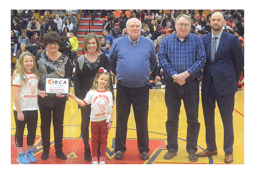 Yorkville resident Becky Hejtmanek, second from left, daughter of the late West Aurora High School boys basketball head coach John McDougal, shows an Illinois Basketball Coaches Association (IBCA) plaque Friday which honors Blackhawk teams for surpassing 1,500 victories. Foreground is Beatrice Clark, 5, Oswego, McDougal great-granddaughter; back row from left are Juliet Clark, 9, Oswego, McDougal great-granddaughter; Hejtmanek; Mary Schroeder, St. Charles, McDougal daughter; Gordon Kerkman, Aurora, retired Blackhawk head coach (1976-2015); Dave Dorsey, Elburn, son of the late head coach Dick Dorsey (1955-1960); and Brian Johnson, West Aurora head coach. John McDougal was head coach 1965-1976. West Aurora teams had collected 1,608 victories through Saturday’s 57-26 victory against visiting Glenbard South. Al Benson/The Voice